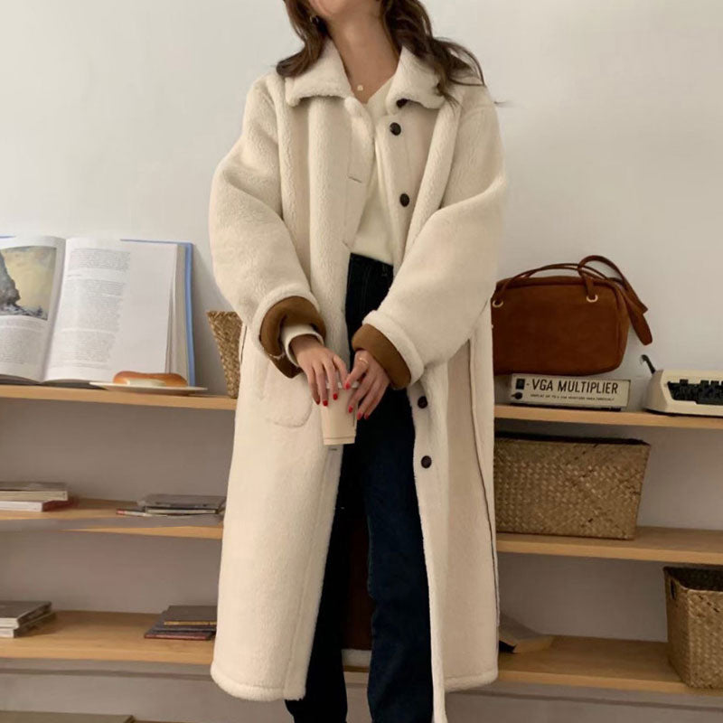 Person wearing a long cream coat with brown cuffs and collar in a room with shelves.