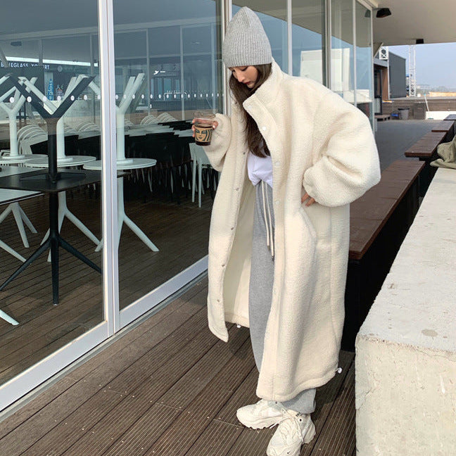 Person wearing a long white coat and gray beanie standing on a wooden deck with glass walls.