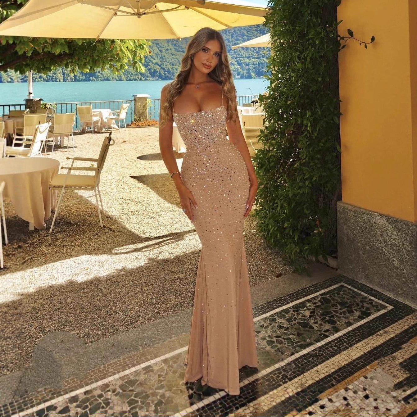 Woman in a beige evening gown standing outdoors with tables and chairs in the background.