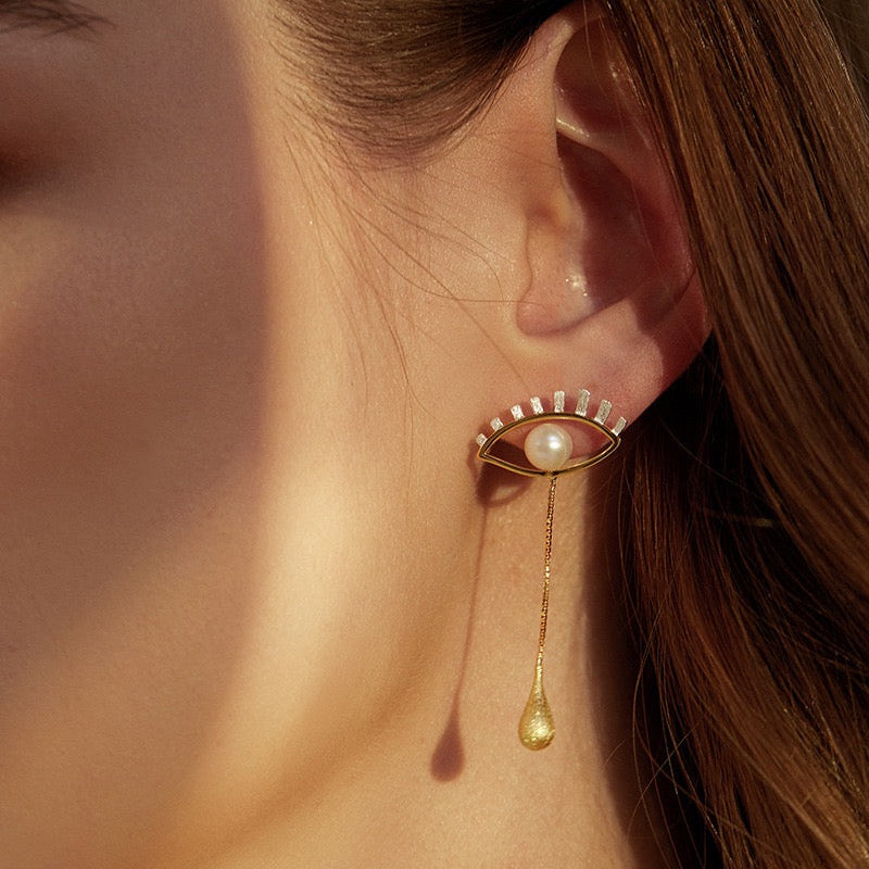 Close-up of a gold earring with a pearl and teardrop pendant worn by a person.