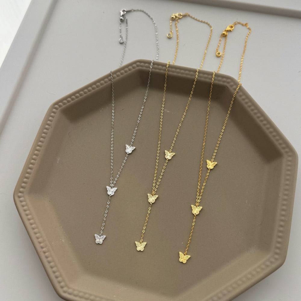 Three butterfly necklaces in silver and gold on a beige tray.
