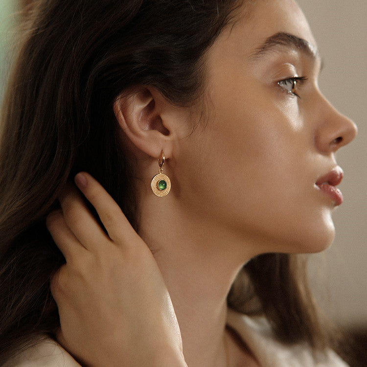 Close-up of a woman wearing gold earrings with a green gemstone, adjusting her hair.