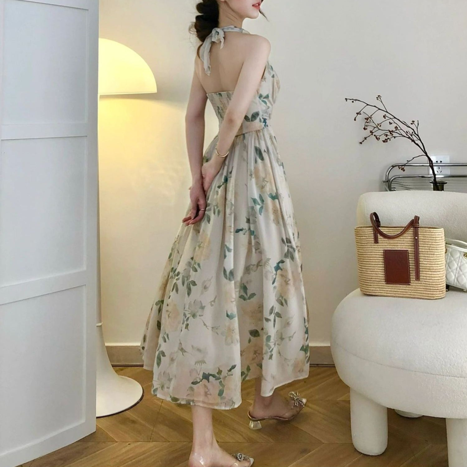 Woman in a floral dress standing in a room with a lamp and table.