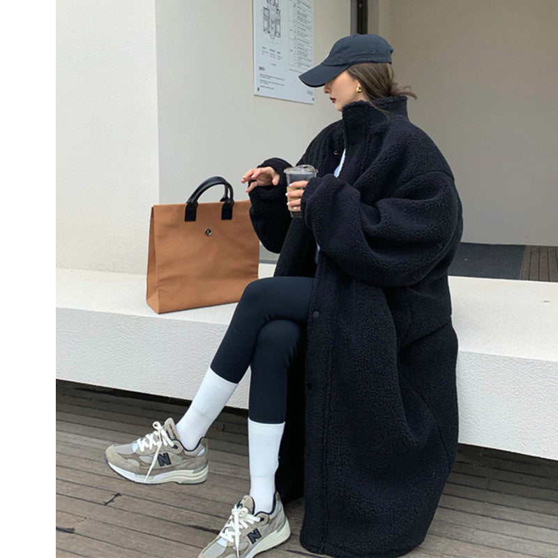 Person sitting on a bench wearing a long black coat, black pants, and a cap, holding a brown bag.