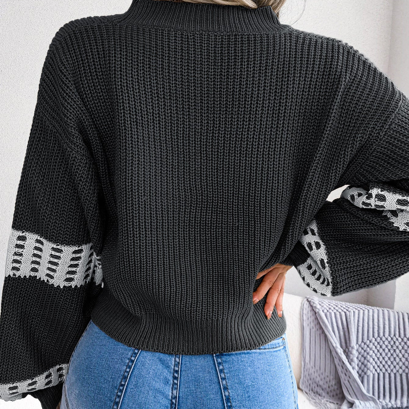 Person wearing a black knit sweater with patterned sleeves and blue jeans indoors.