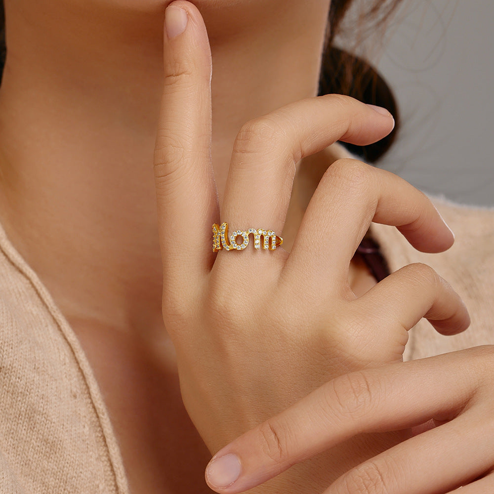 Gold ring with 'mom' on a person's finger against a neutral background