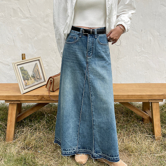 Person wearing a long denim skirt standing next to a wooden bench with a framed picture and handbag.
