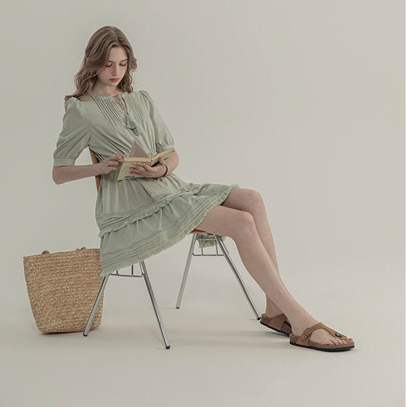 Woman in a green dress sitting on a chair with a woven bag beside her against a plain background