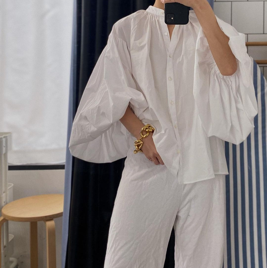 Person wearing a white outfit taking a mirror selfie in a room with curtains and a table.