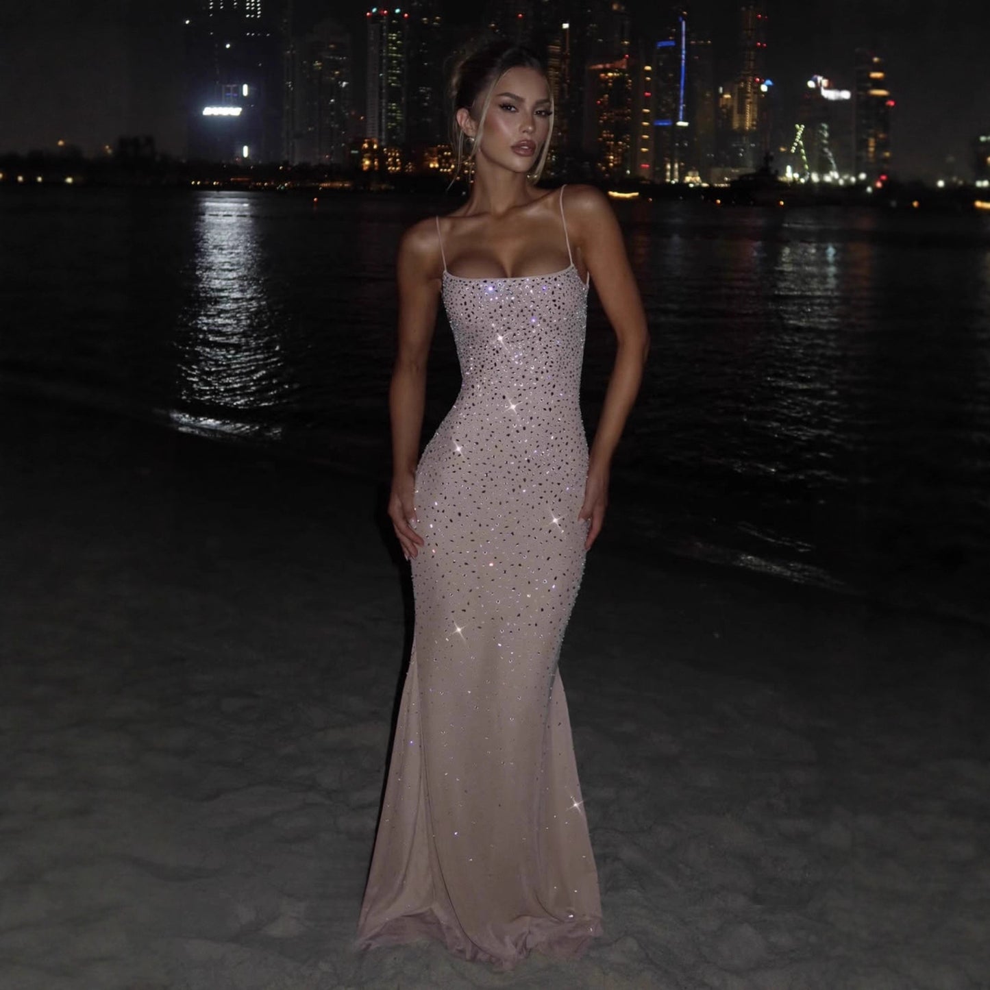 Woman in a sparkling evening gown standing on a beach at night with city lights in the background
