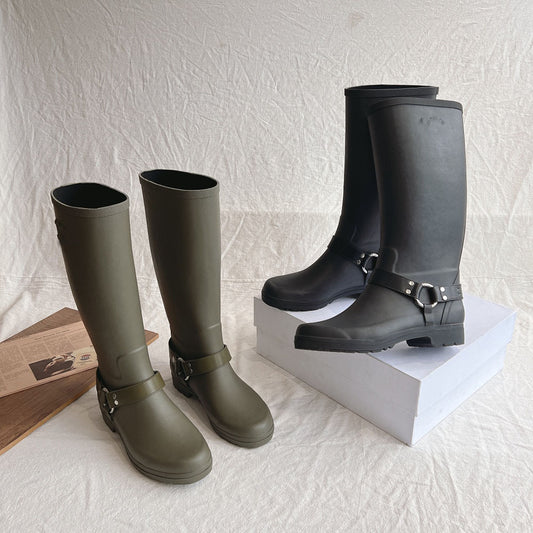 Two pairs of rain boots, one green and one black with buckles, on a textured white surface.