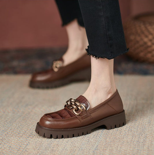 Brown loafers with gold chain detail worn by a person on a textured floor.