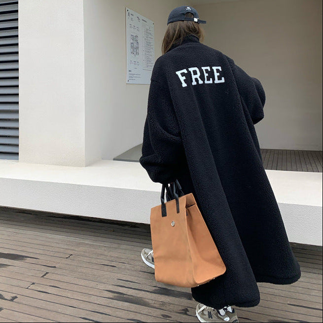Person wearing a black coat with 'FREE' text, holding a tan bag, walking in an indoor setting.