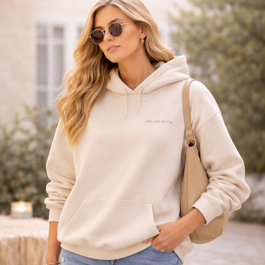 Woman wearing a beige hoodie with 'Malone Boutique' logo, holding a beige handbag outdoors.