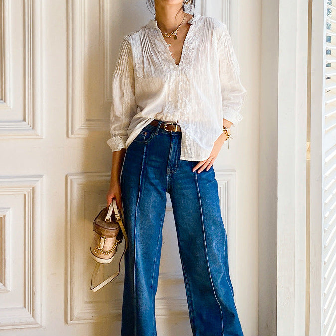 Woman wearing a white blouse, blue jeans, and a hat indoors.