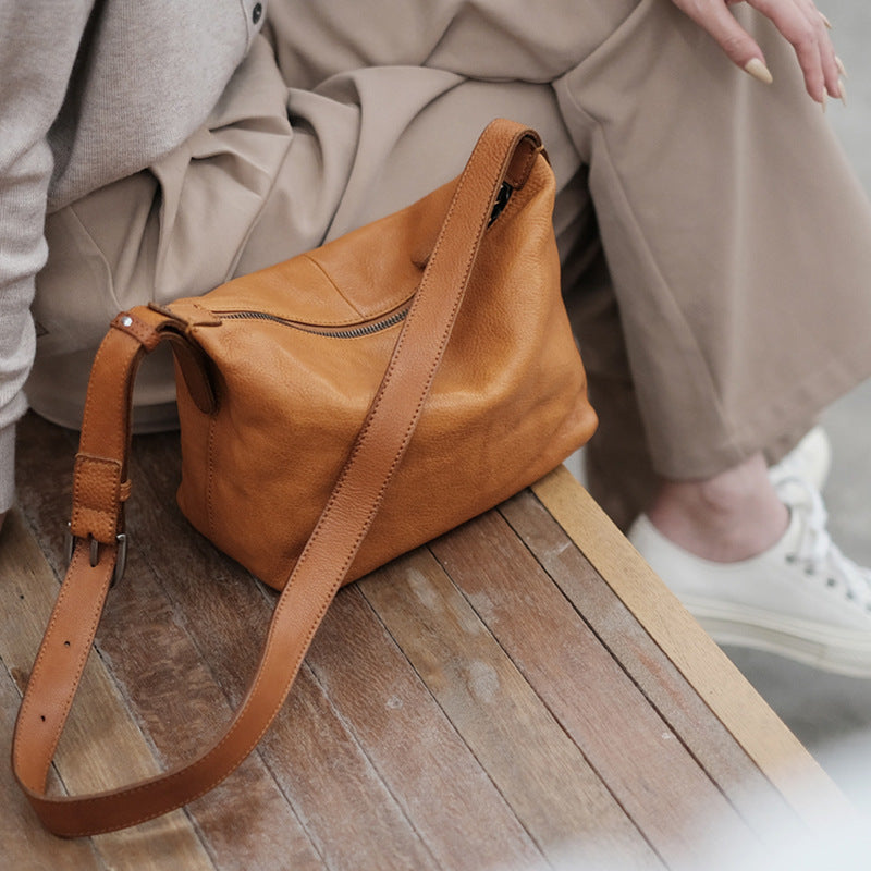 Brown leather handbag on a wooden surface with a person wearing beige pants in the background.