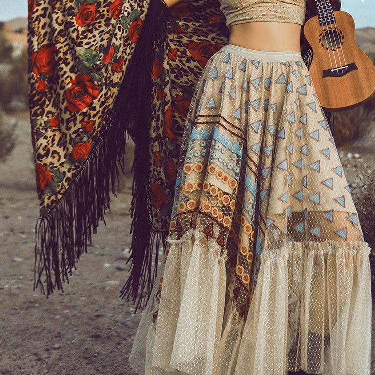 Person wearing a patterned skirt and leopard print shawl with a guitar, standing on a dirt path.