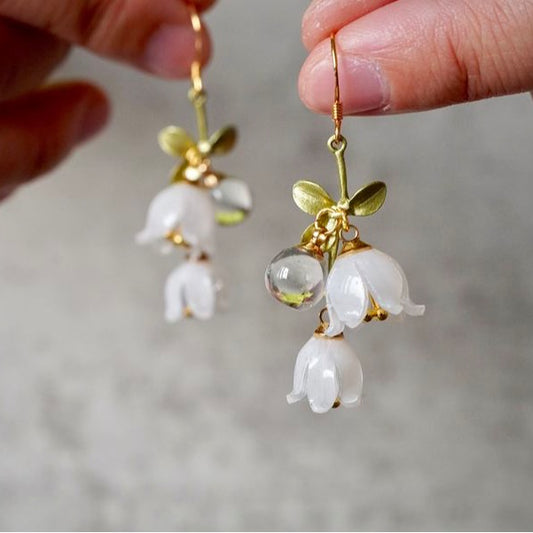 Gold earrings with white floral elements held by a hand against a neutral background