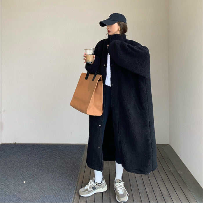 Person wearing a black coat and cap, holding a brown bag and cup, standing against a plain wall.