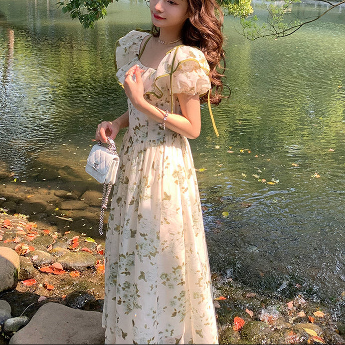 Woman in a floral dress standing by a lake with trees and a bridge in the background