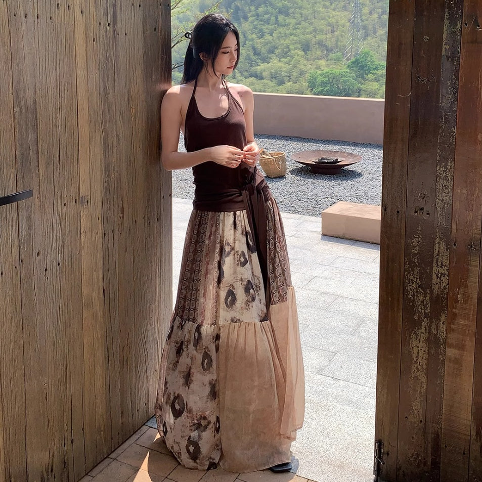 Woman in a long, patterned dress standing in a sunlit outdoor area with wooden walls.