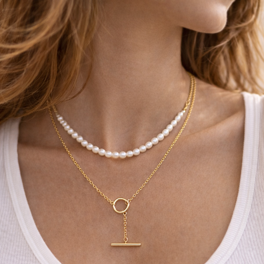 Gold necklace with a pendant and pearl necklace on a person wearing a white tank top.